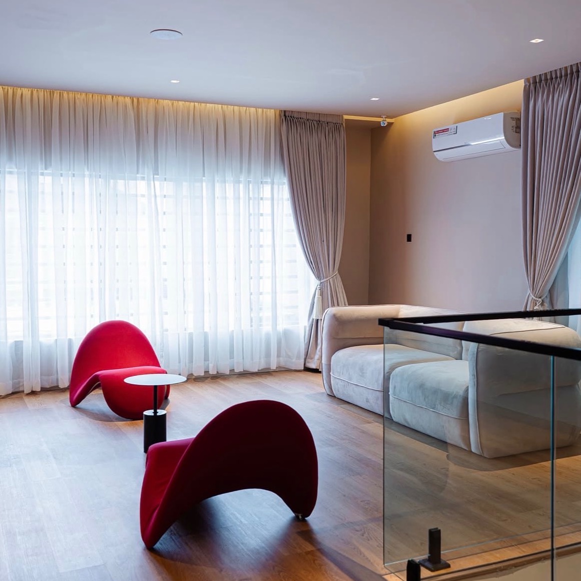 A bright living space with sculptural crimson lounge chairs, sheer drapery and warm wood floors by Bestchoice Interiors