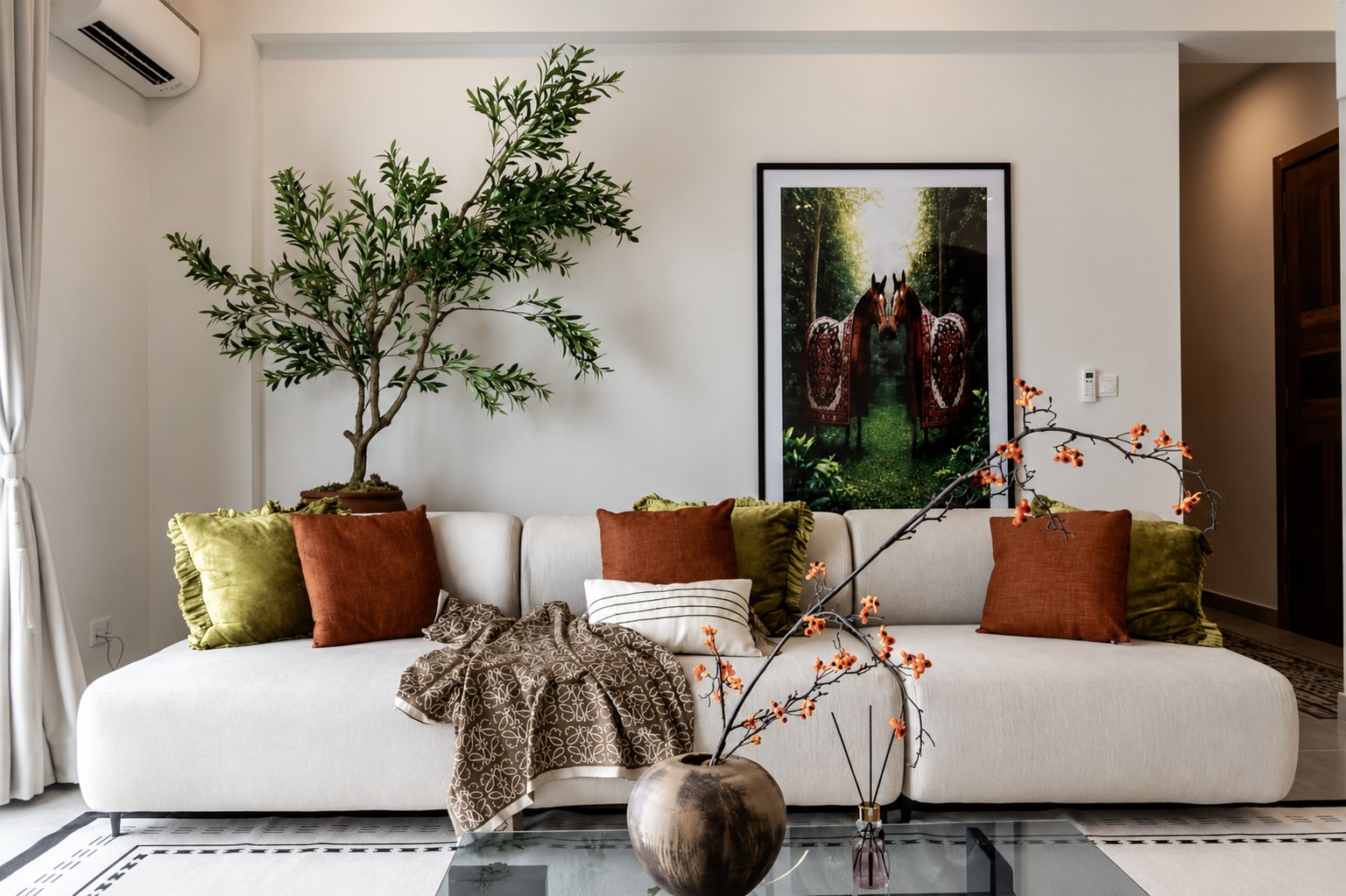 A styled living room by Luxxterior — cream sofa, olive and terracotta cushions, framed art and a sculptural olive tree.